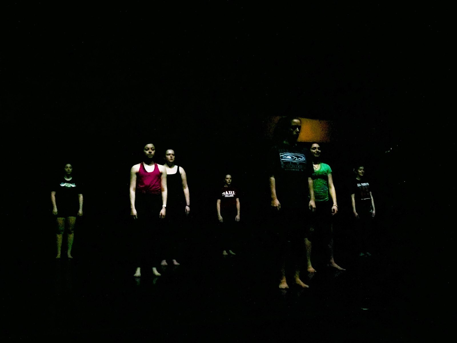 A line of dancers standing in low light, faces and torsos catching a faint glow